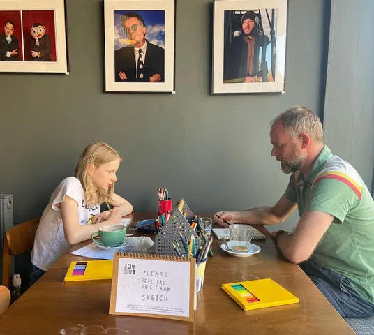 A father and daughter drawing together at Art Club, with coloured pencils and coffee on the table